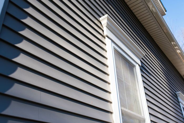 Gray horizontal siding on a house, white trim around a window, blue sky.