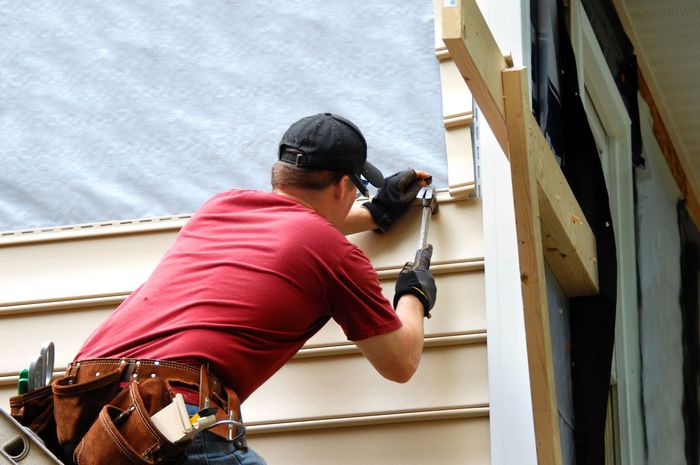 Person in a red shirt installing siding on a building's exterior, using a hammer and wearing a tool belt.