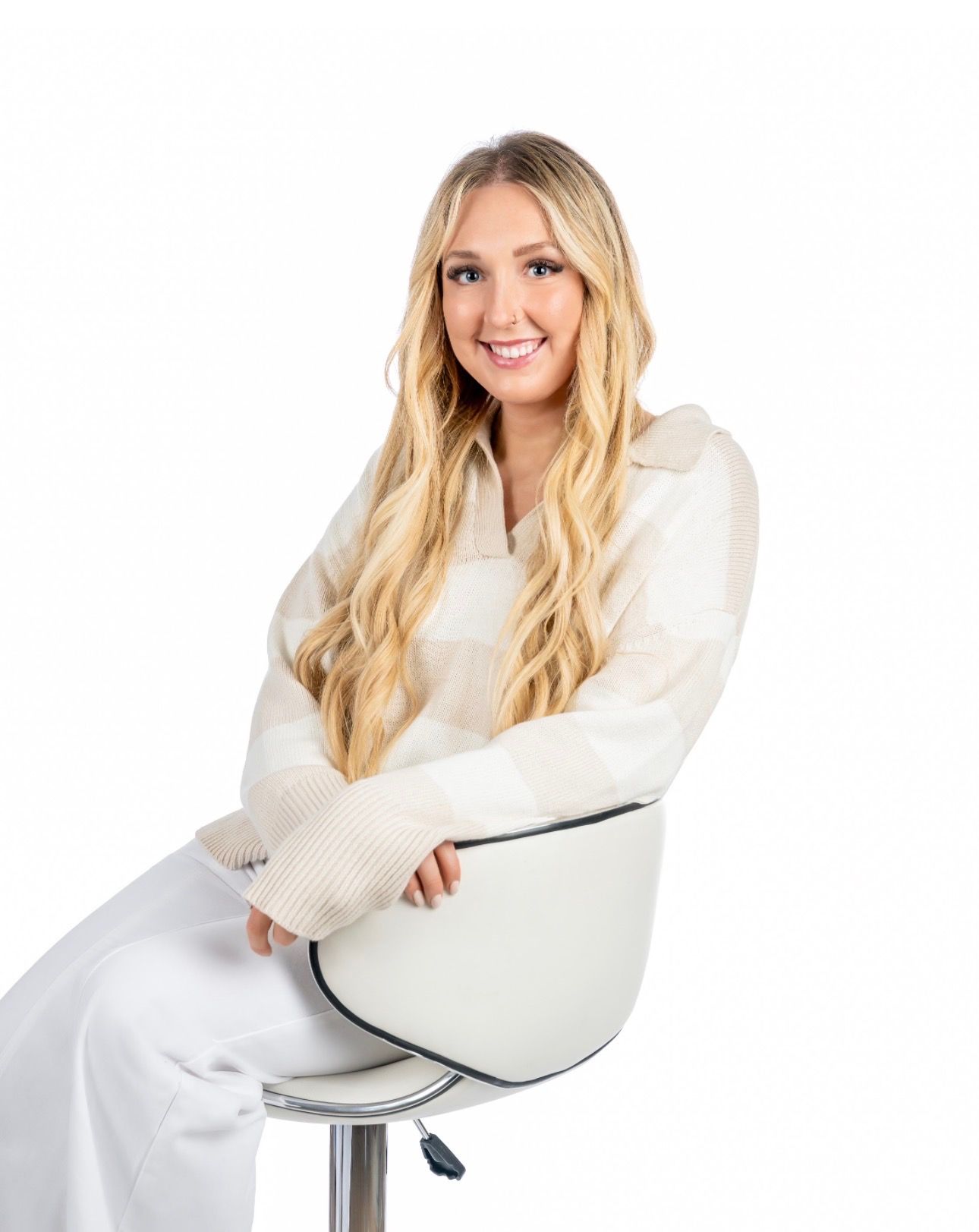 Woman with long blonde hair, smiling, seated on a white chair, wearing a cream sweater and white pants.