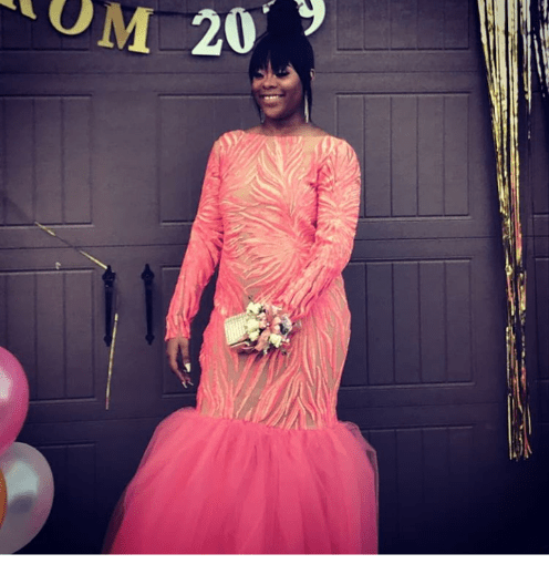 Person in a coral gown holding flowers, standing in front of a decorated backdrop at a prom event