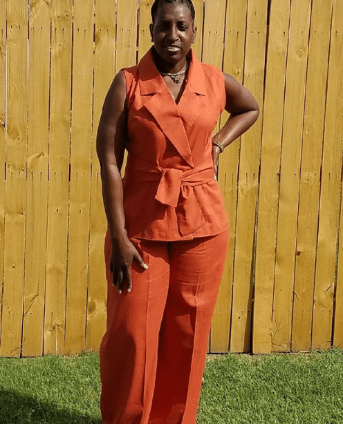 Person in an orange outfit standing on grass in front of a wooden fence, hands on hips.