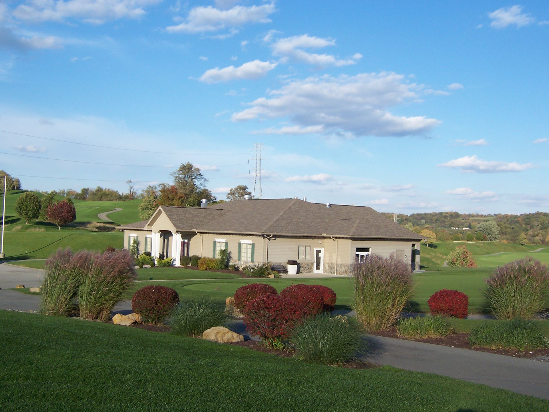Low building with a brown roof and a green lawn, shrubs, and trees under a blue sky with clouds.
