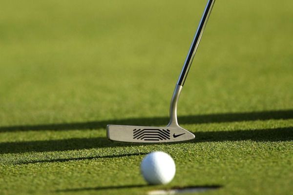 Person in red shirt and khaki shorts putting golf ball on a green. Others in the background.