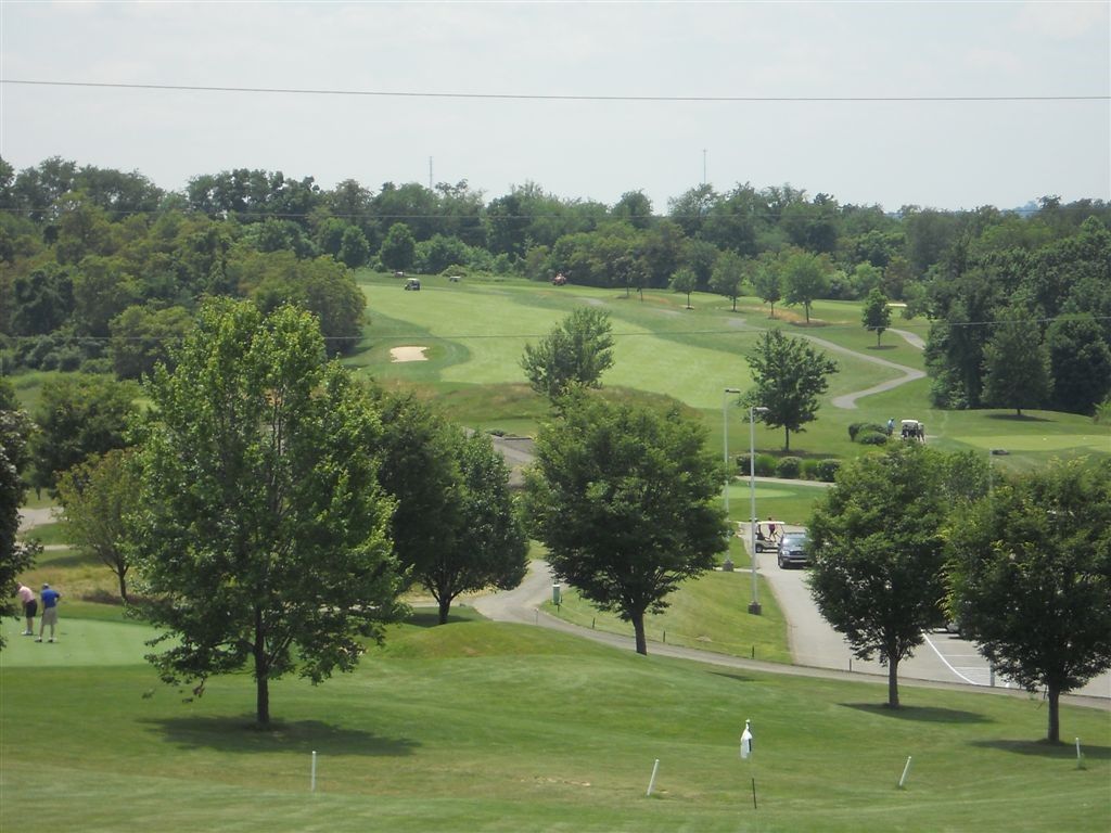 Golf course view, green fairways and trees, players on the green, golf carts on path, sunny day.