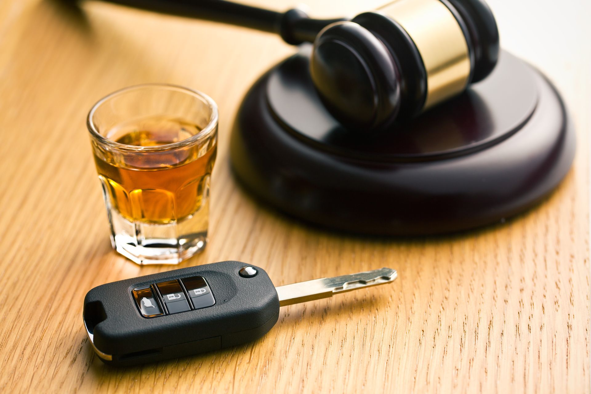 Shot glass with amber liquid, car keys, and gavel on a wooden surface.