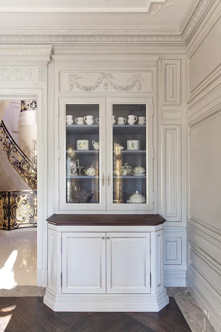A white cabinet with glass doors is in a room next to a staircase.