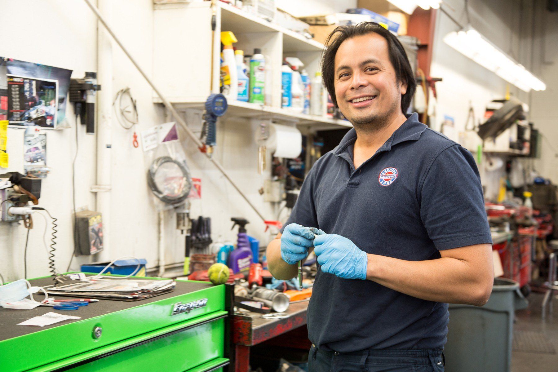 Mechanic in blue shirt and gloves smiles in his workshop, holding a part, with tools and shelves in the background.