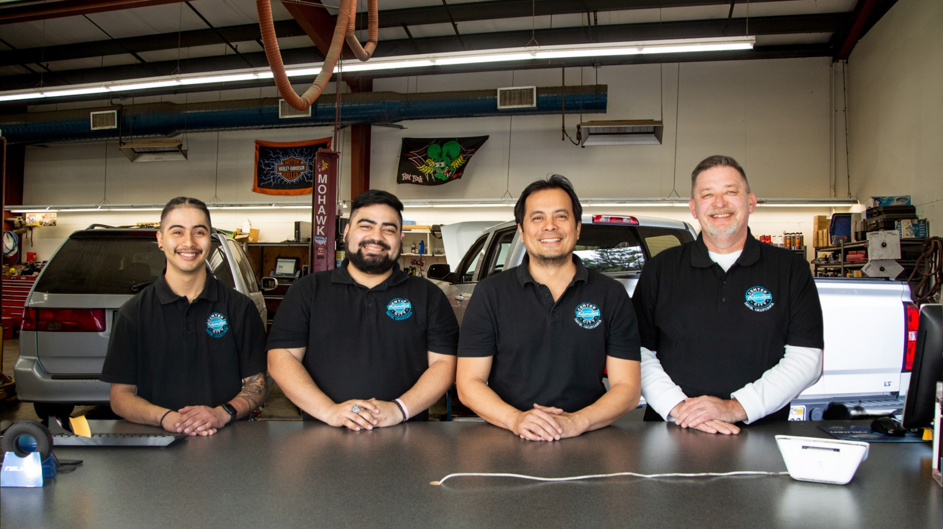 Four men in black shirts pose behind a counter in an auto repair shop.