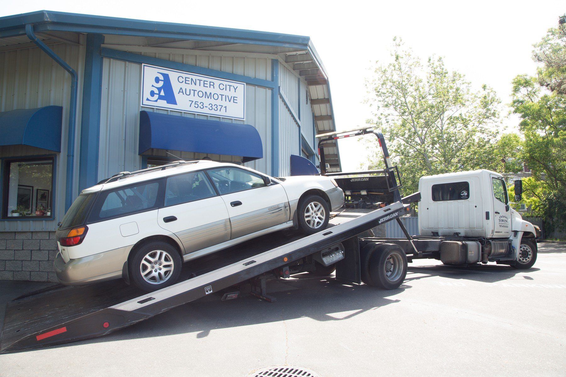 White station wagon on a flatbed tow truck in front of a blue and white auto repair shop.