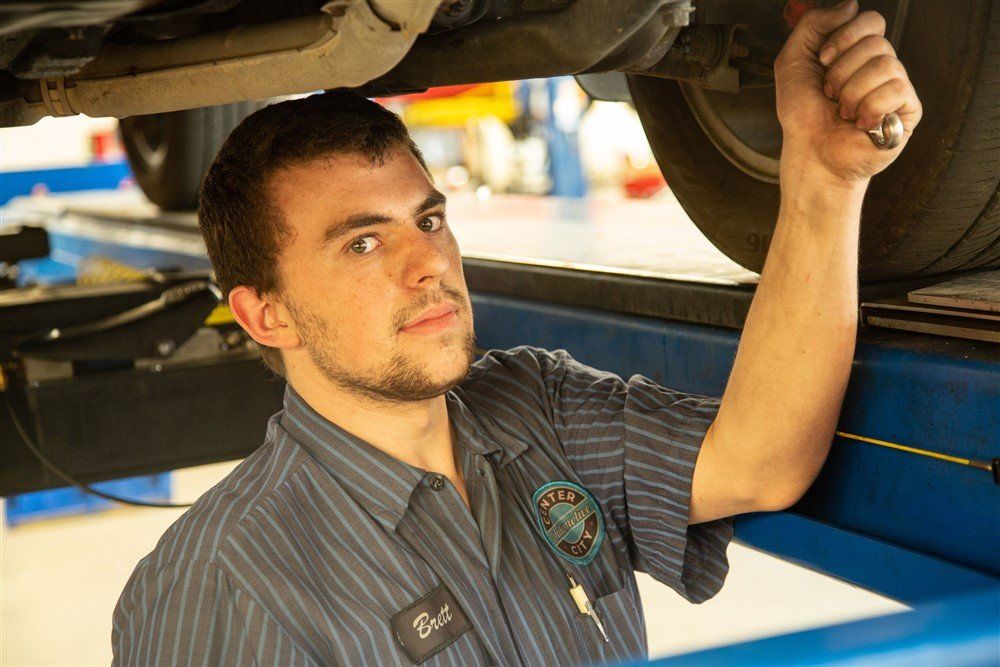 Mechanic working on a car, holding a wrench under the vehicle, blue uniform, looking toward the viewer.