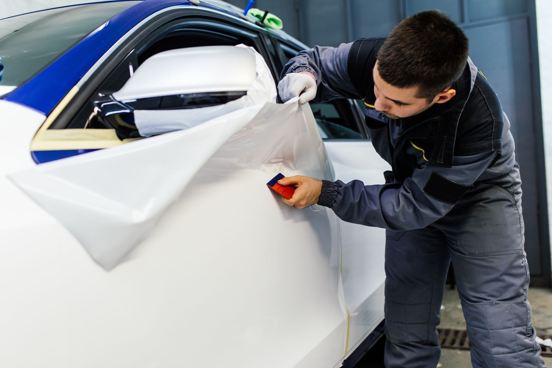 A person in a work suit applies white vinyl wrap to a car door in a garage, using a squeegee.