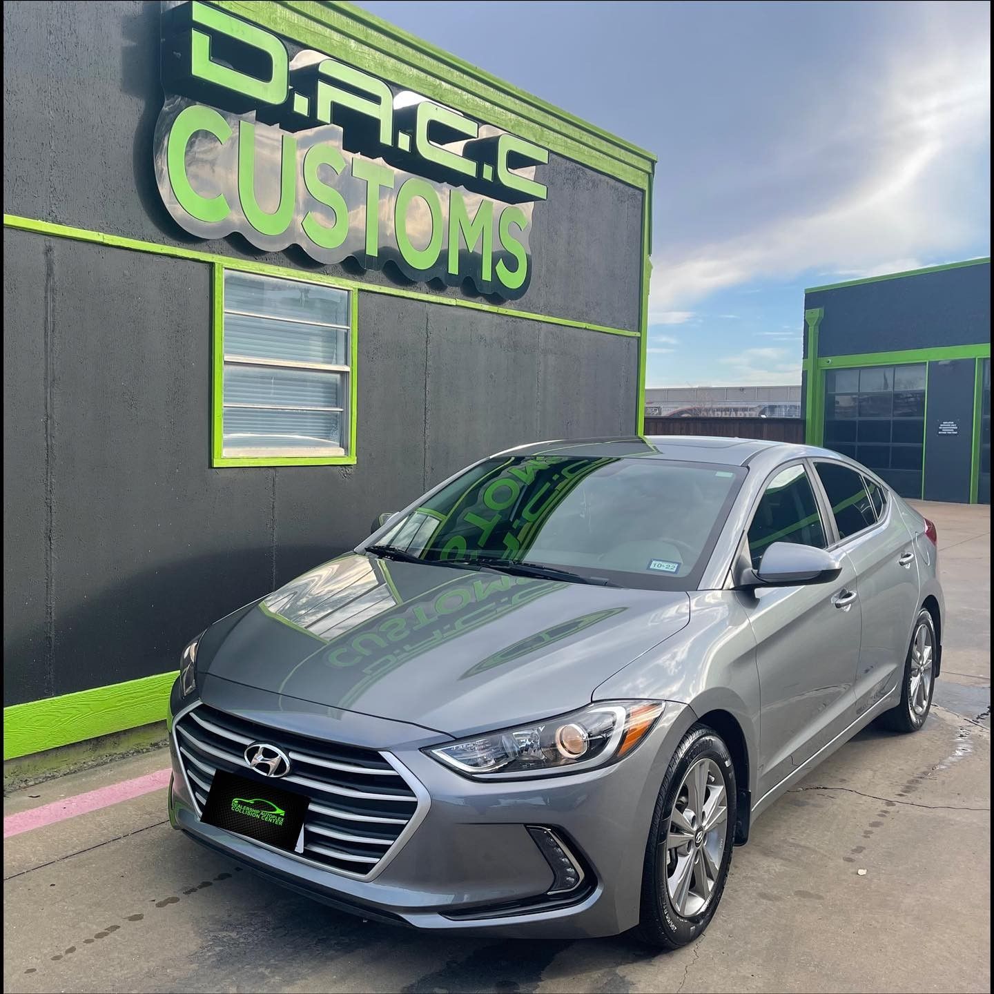A silver Hyundai Elantra parked in front of a custom auto shop with a green and black facade, under a cloudy sky.