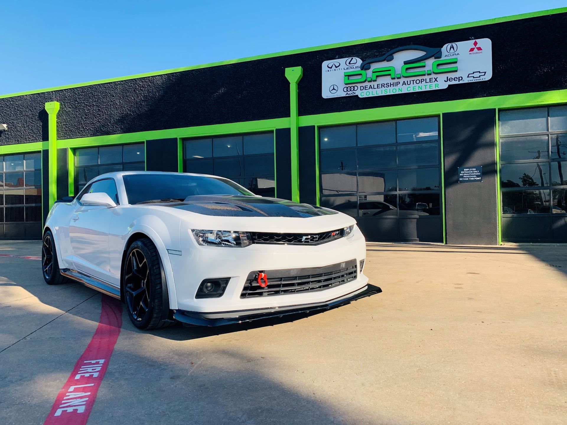 White Camaro with black accents parked in front of a green and black building. Red tow hook visible.