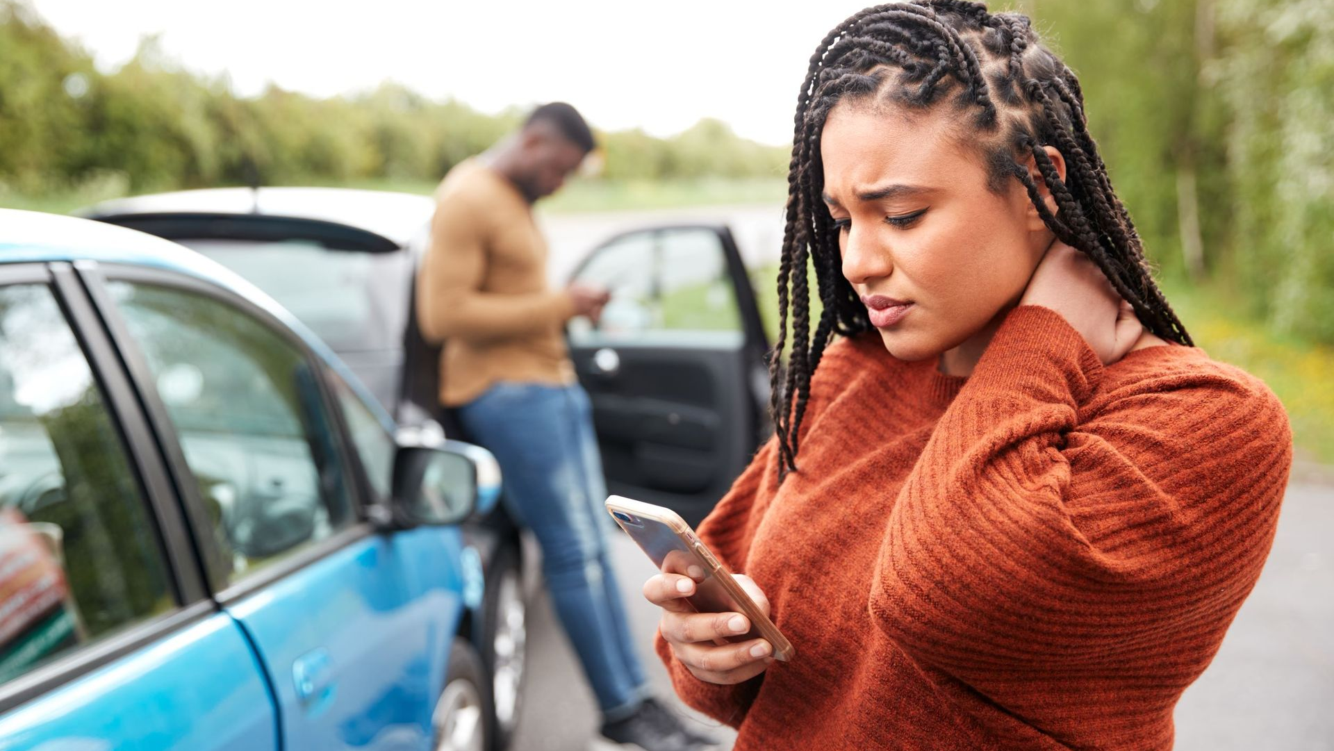 Woman holding neck, looking at phone, near damaged blue car. Man near the car on his phone.