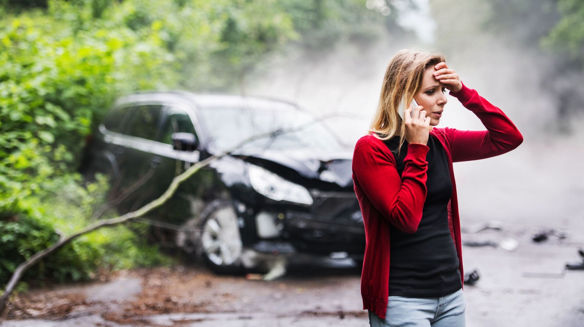 Woman in red cardigan on phone, standing near a damaged black car after a car accident on a road.