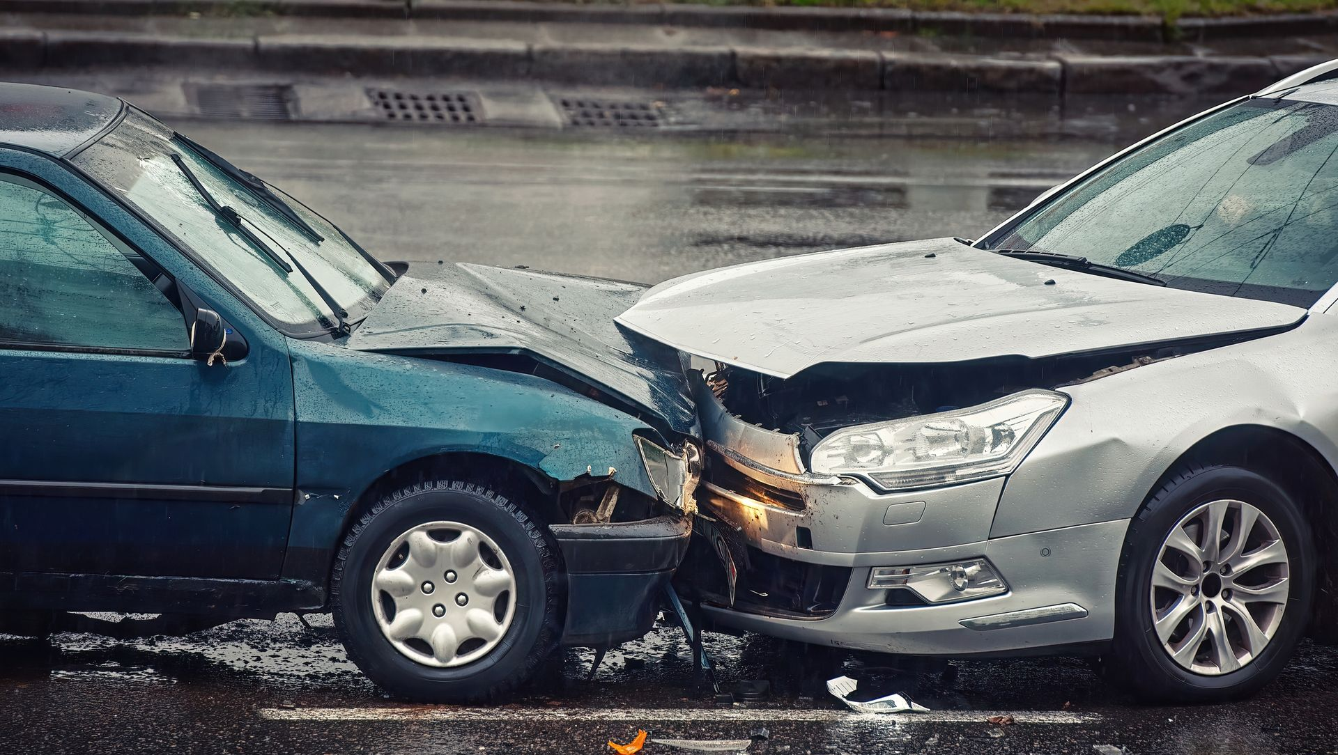 Two cars, teal and silver, crashed on a wet street. Both front ends are heavily damaged.
