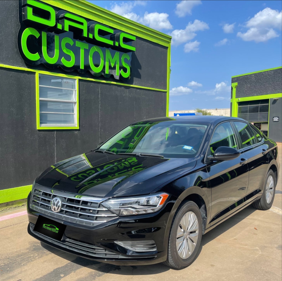 A black Volkswagen Jetta parked outside a custom shop with a green and black sign that reads 