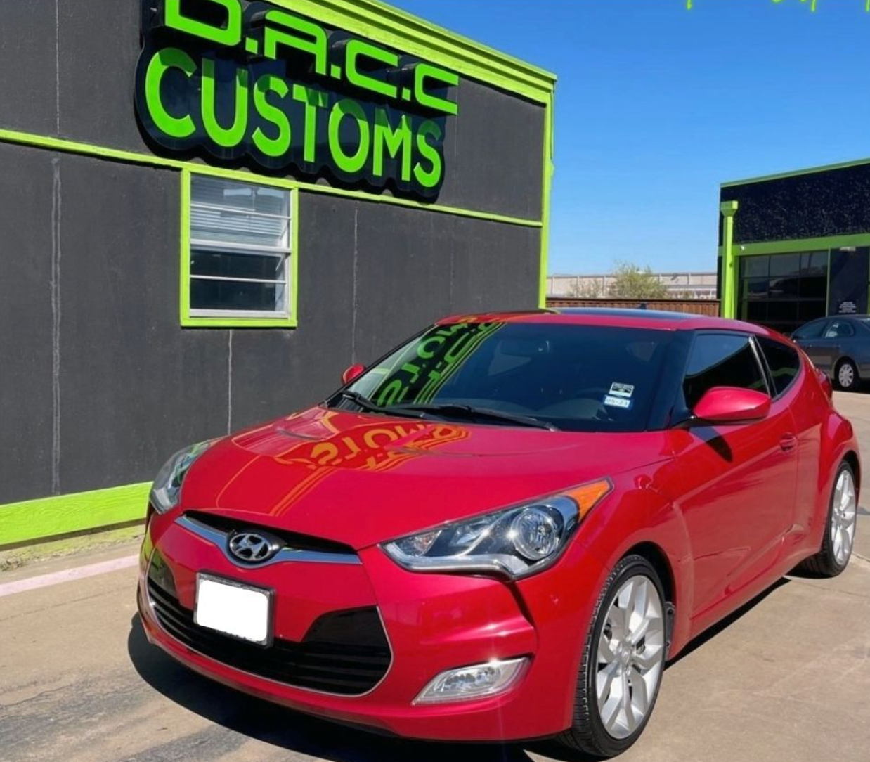 Red Hyundai Veloster parked in front of a D.A.C.C. Customs shop, on a sunny day.