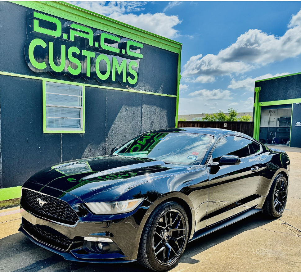 Black Ford Mustang parked in front of D.A.C.C. Customs building with green trim.