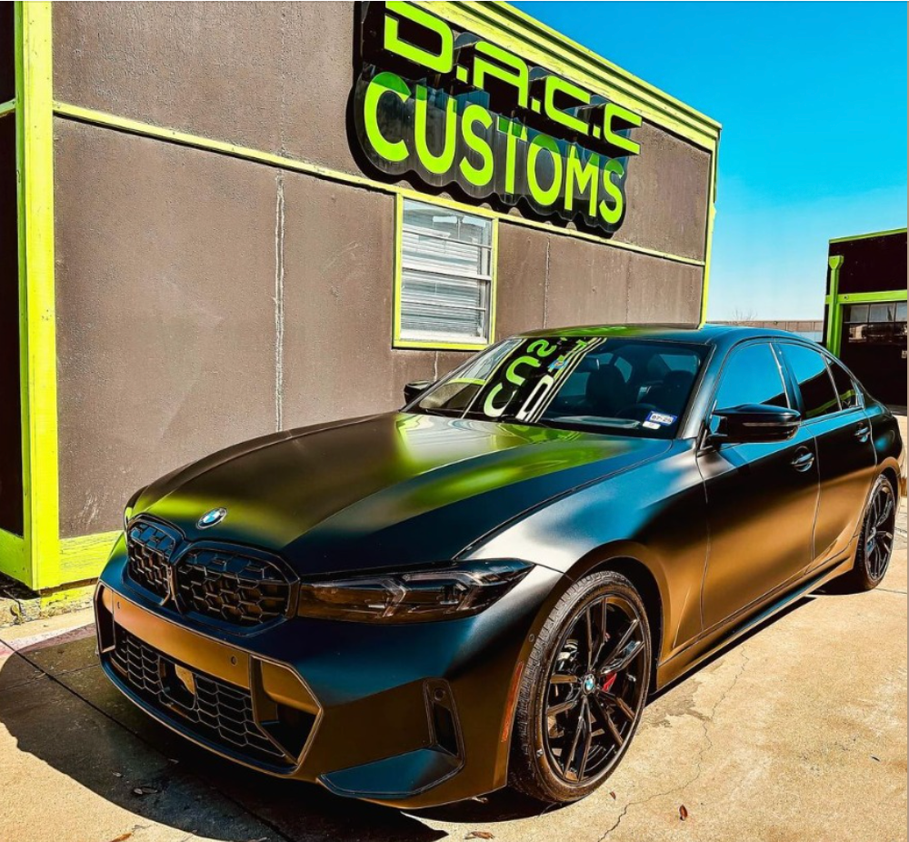 Black BMW sedan parked in front of a shop called D.R.C. Customs with green and black accents. The car has black rims and a matte finish.