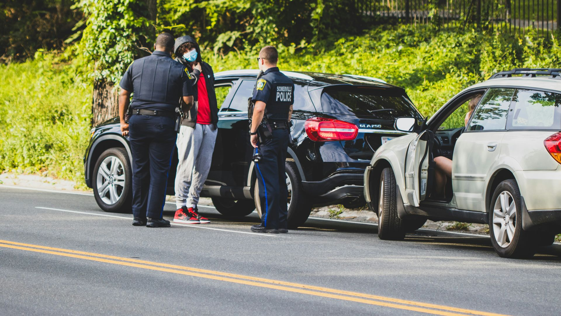 Police officers interacting with a person near two cars parked on a road. One car door is open.