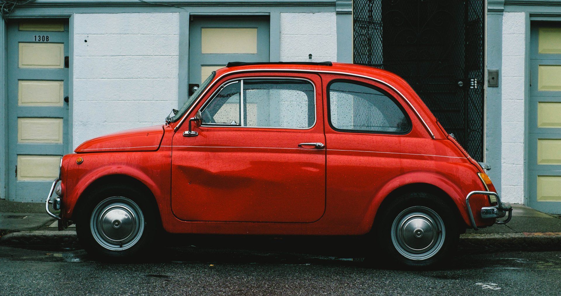 Red vintage Fiat 500 parked on a wet street in front of a building with beige doors.