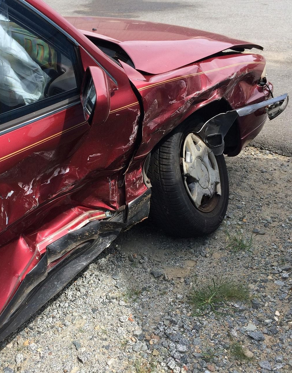 Red car with extensive front-end damage, including deployed airbag, on gravel.