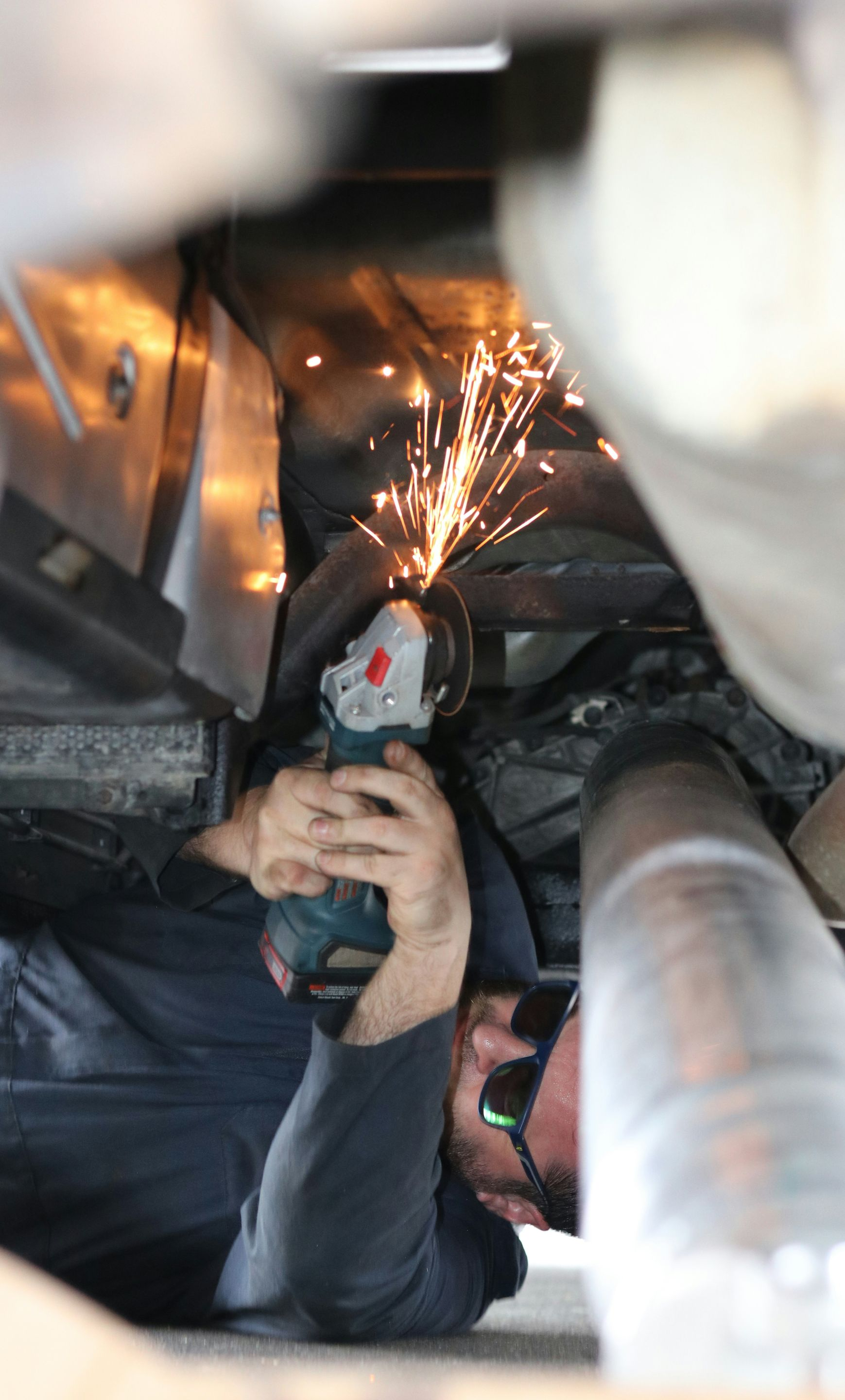 A person using an angle grinder under a vehicle, creating a spray of sparks.