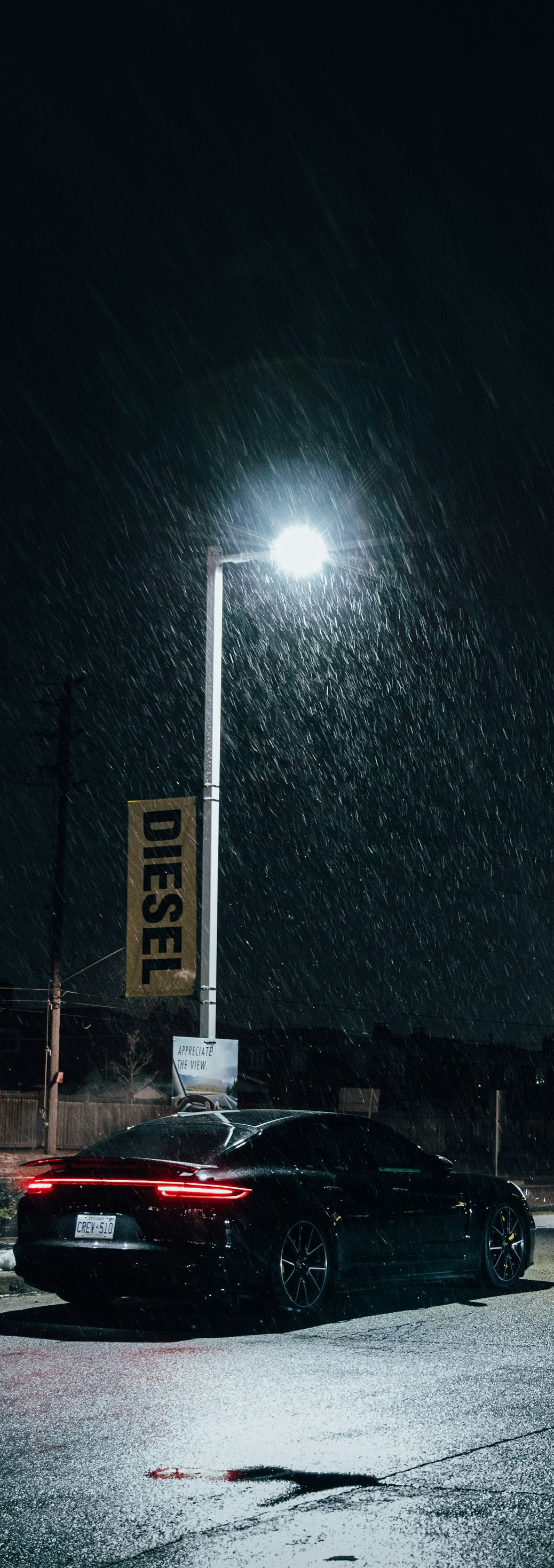 A black car parked under a street lamp at night. It is raining and the diesel sign is visible.