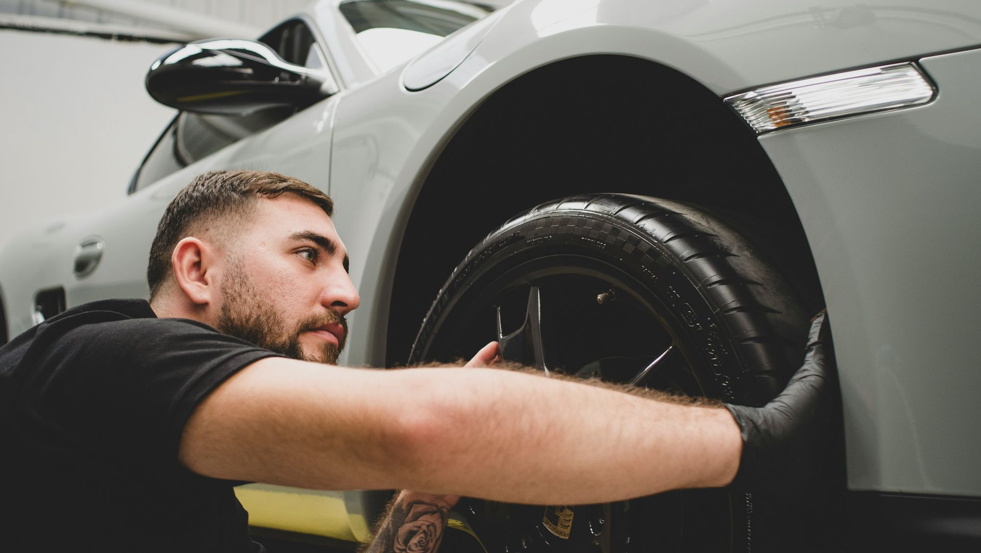 A person wearing a black shirt and gloves works on the wheel of a light gray car inside a garage.