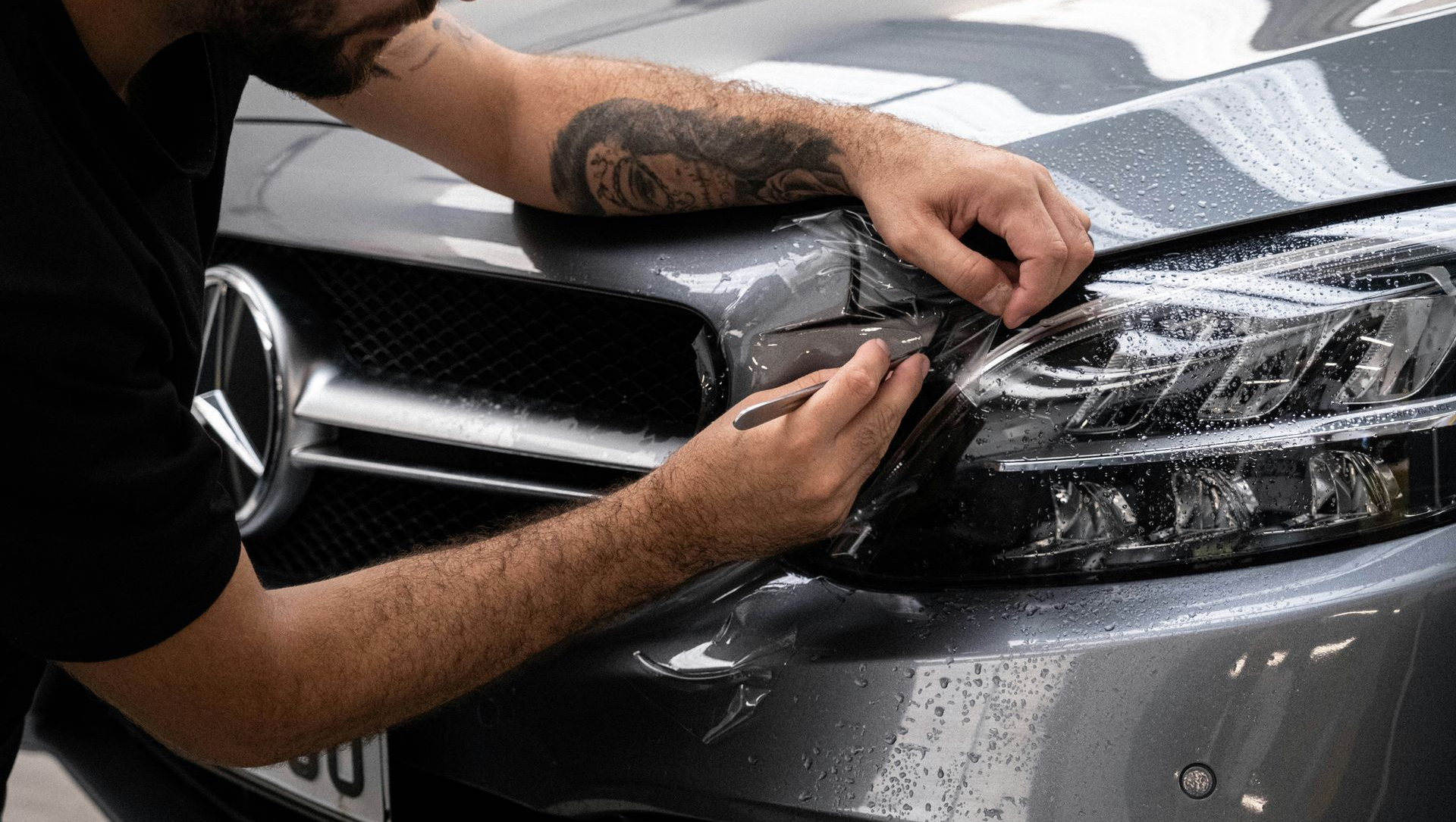 A person carefully applies a protective film to the front fender of a grey Mercedes-Benz using a small tool.