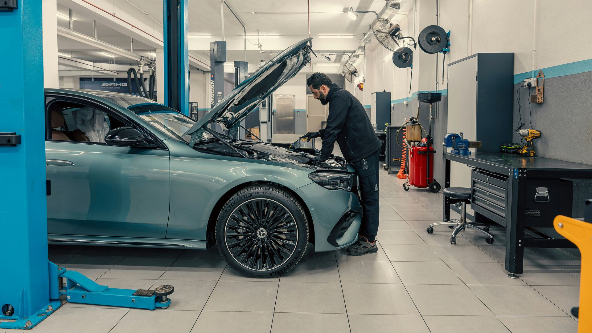A mechanic works under the open hood of a teal luxury sedan inside a bright, well-equipped automotive repair shop.