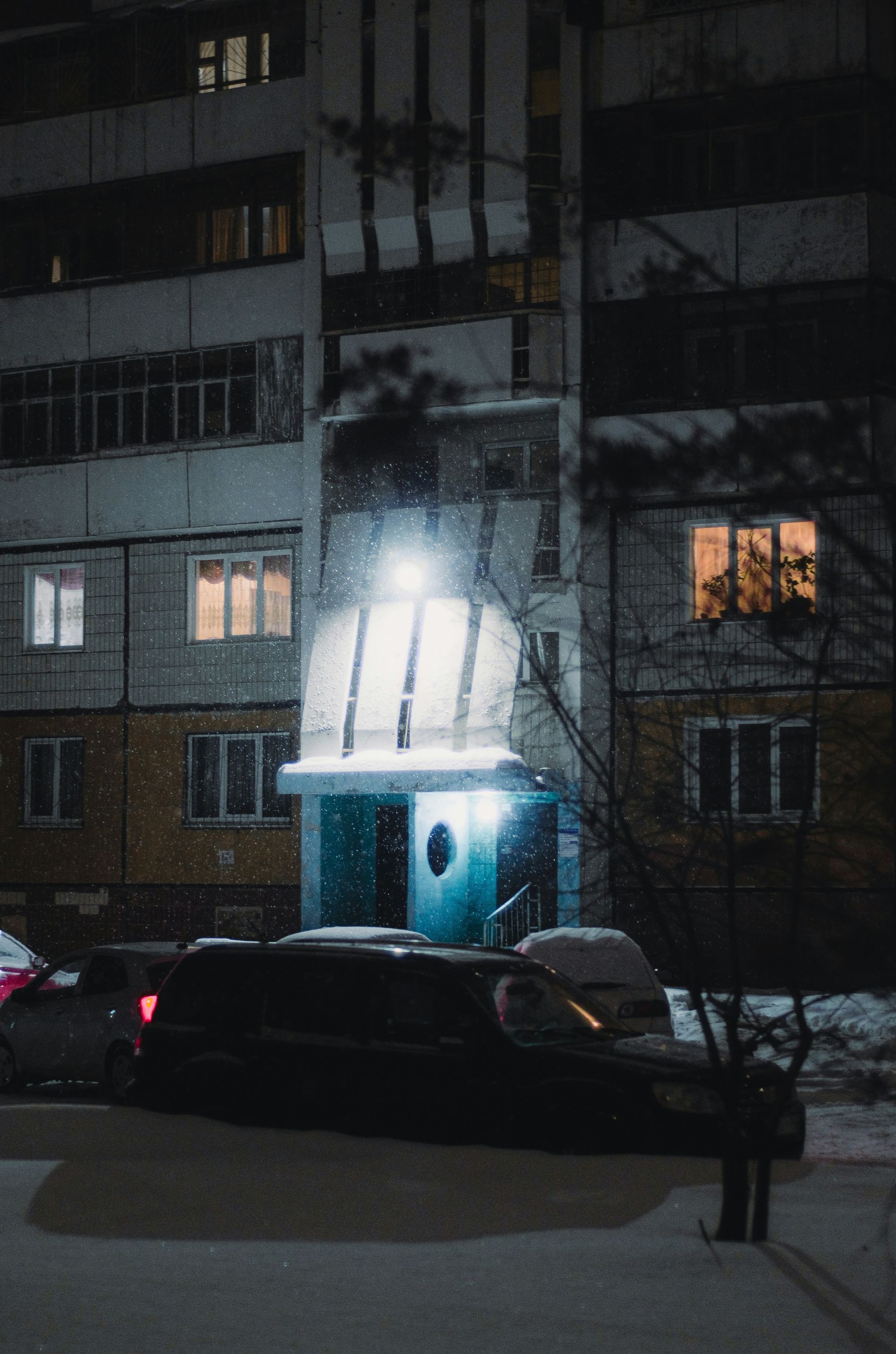 Snowy night scene of a building with lit windows and entrance, car parked in front.