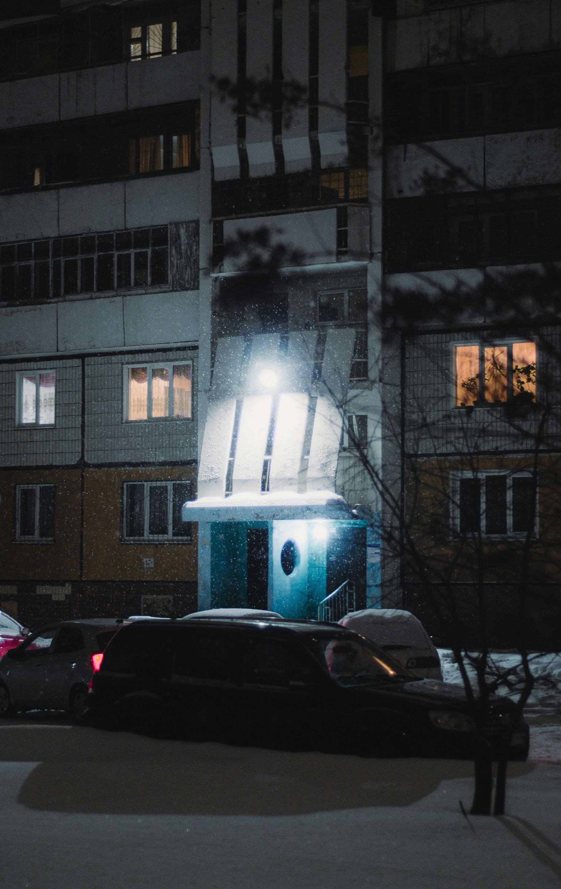 Snowy night scene of a building with lit windows and entrance, a parked car, and a person on the sidewalk.