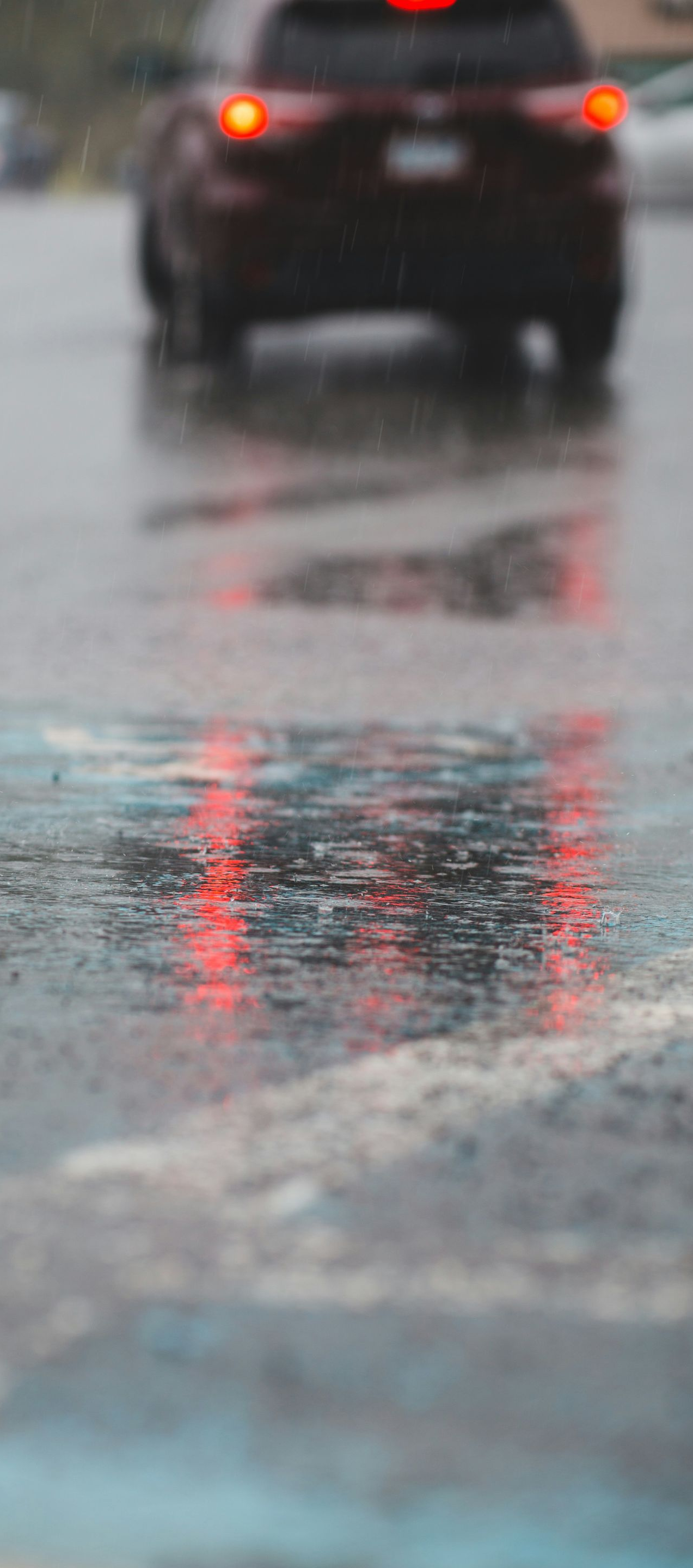 Red taillights reflecting in a wet street, with a dark car driving in the rain.