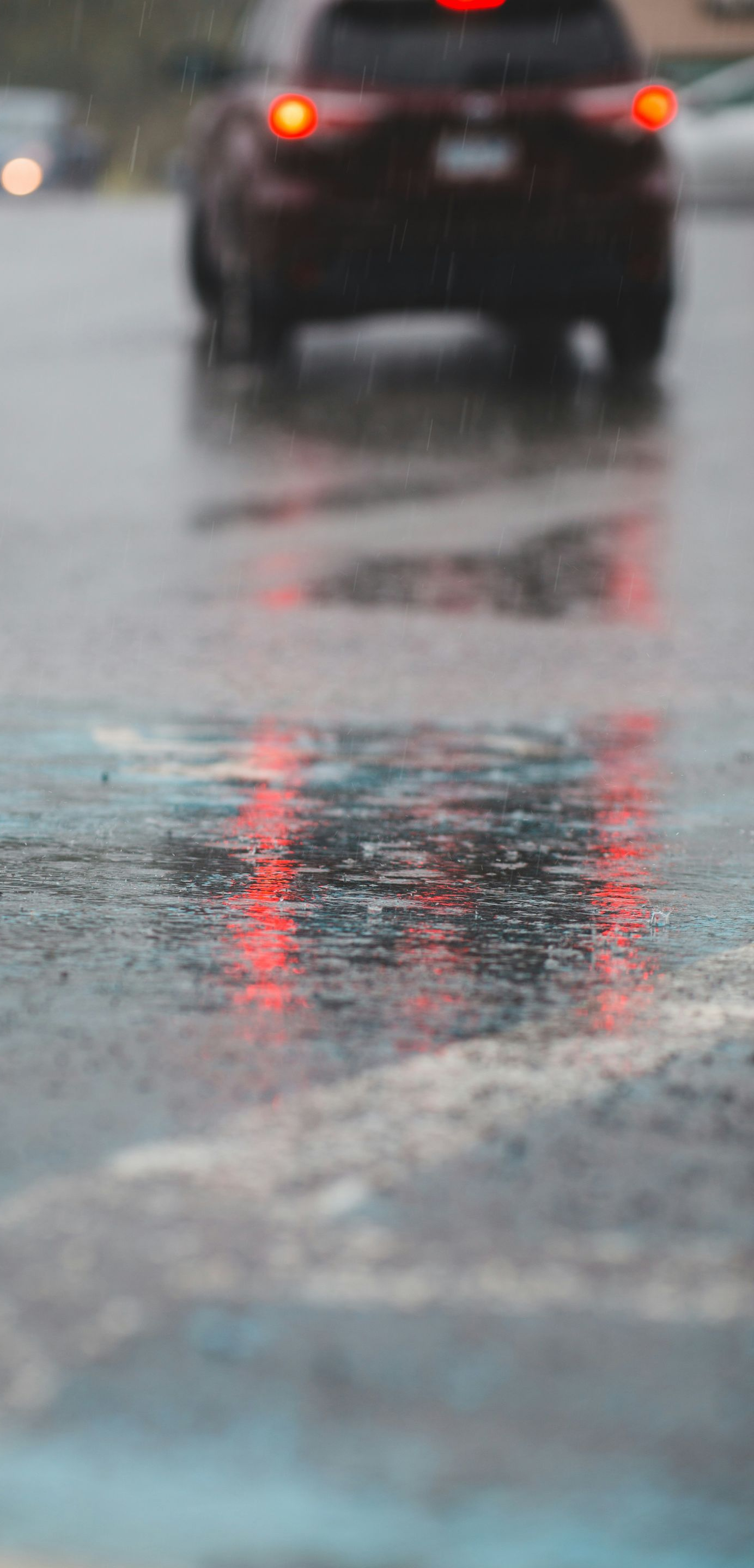 Rainy street scene with red taillights reflecting in a puddle; another car in the distance.