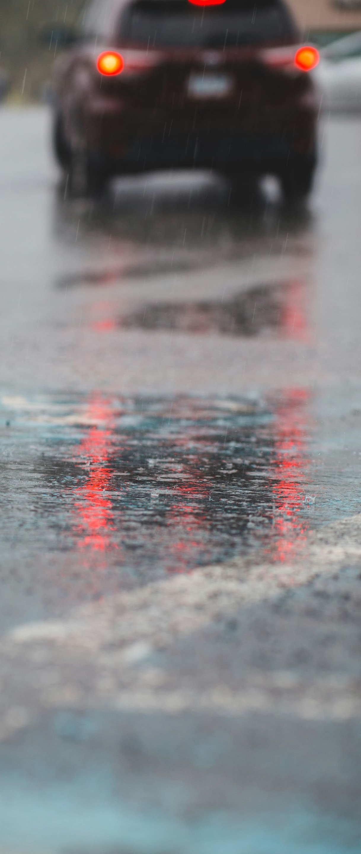 Rainy street scene with red taillights reflecting in a puddle; another car in the distance.