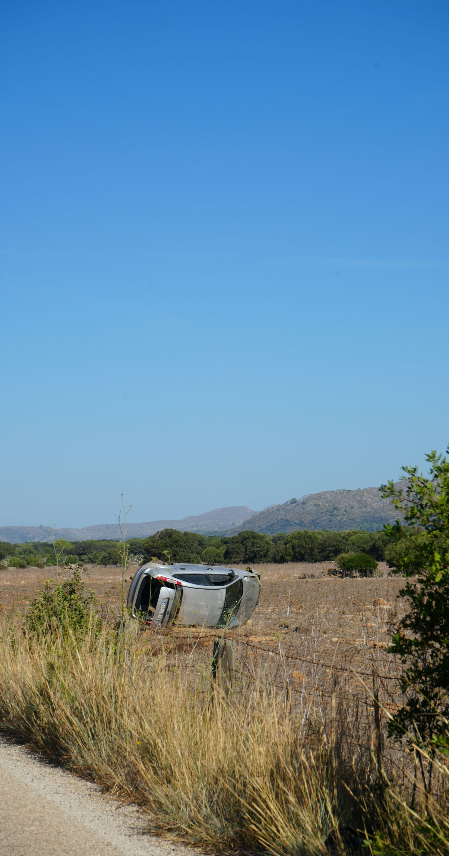Car overturned in a field next to a dirt road under a clear blue sky.