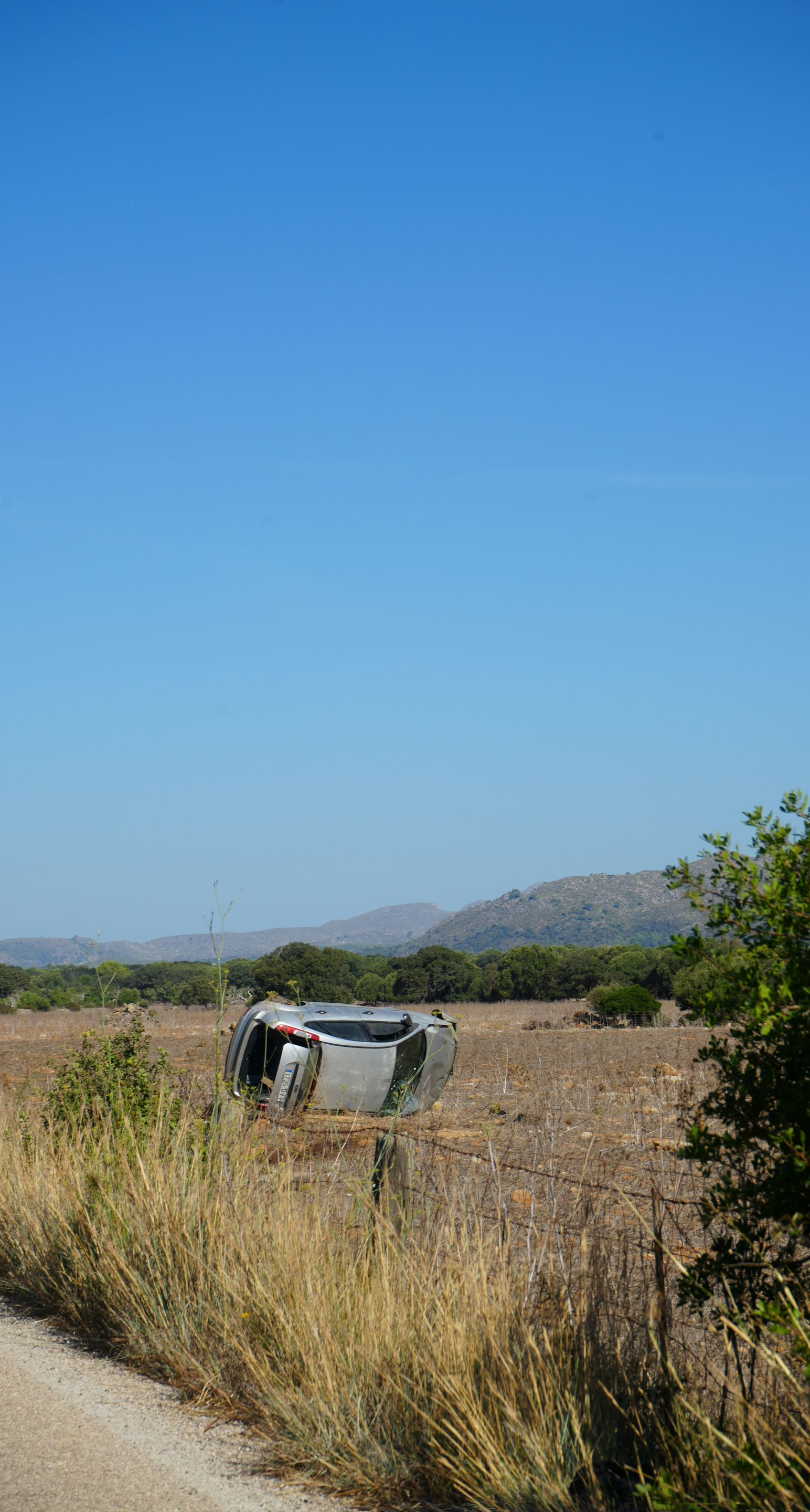 A silver car flipped upside down in a dry, rural field next to a dirt road under a clear blue sky.