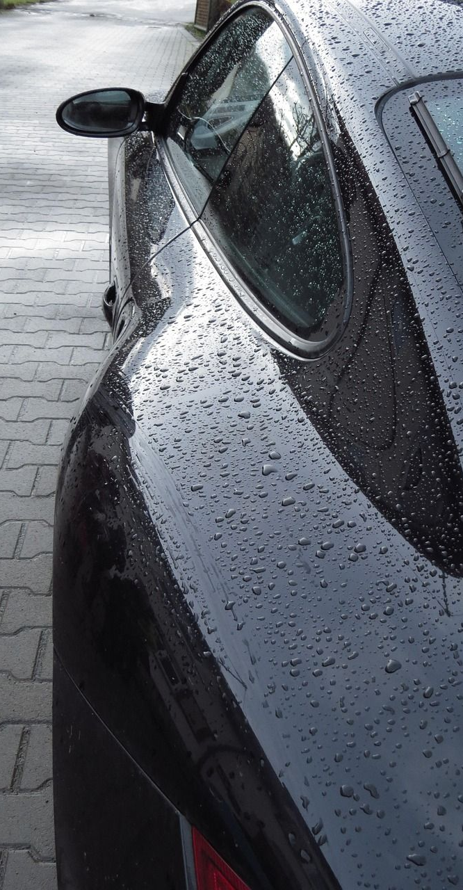 Black sports car, wet with raindrops, parked on a brick-paved driveway.