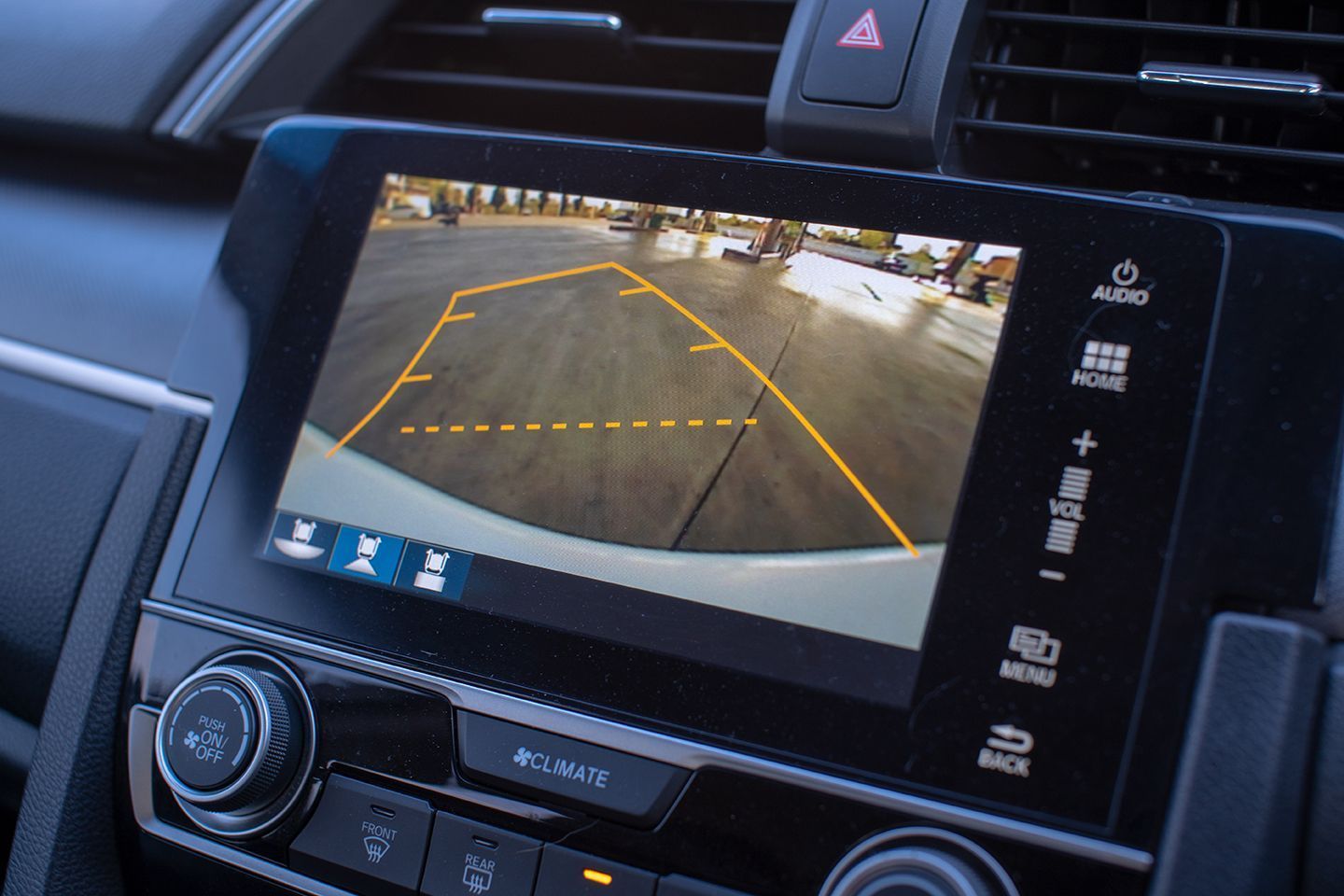 A car's infotainment screen displays a rearview camera view, showing a parking space with guidance lines.