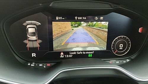 Car's digital dashboard showing a rear view camera view with parking sensors and prompts, in reverse.