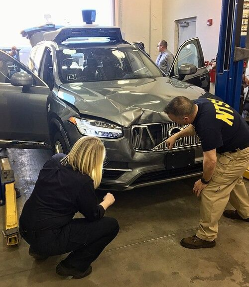 Damaged gray SUV with two people inspecting the front in a garage setting.