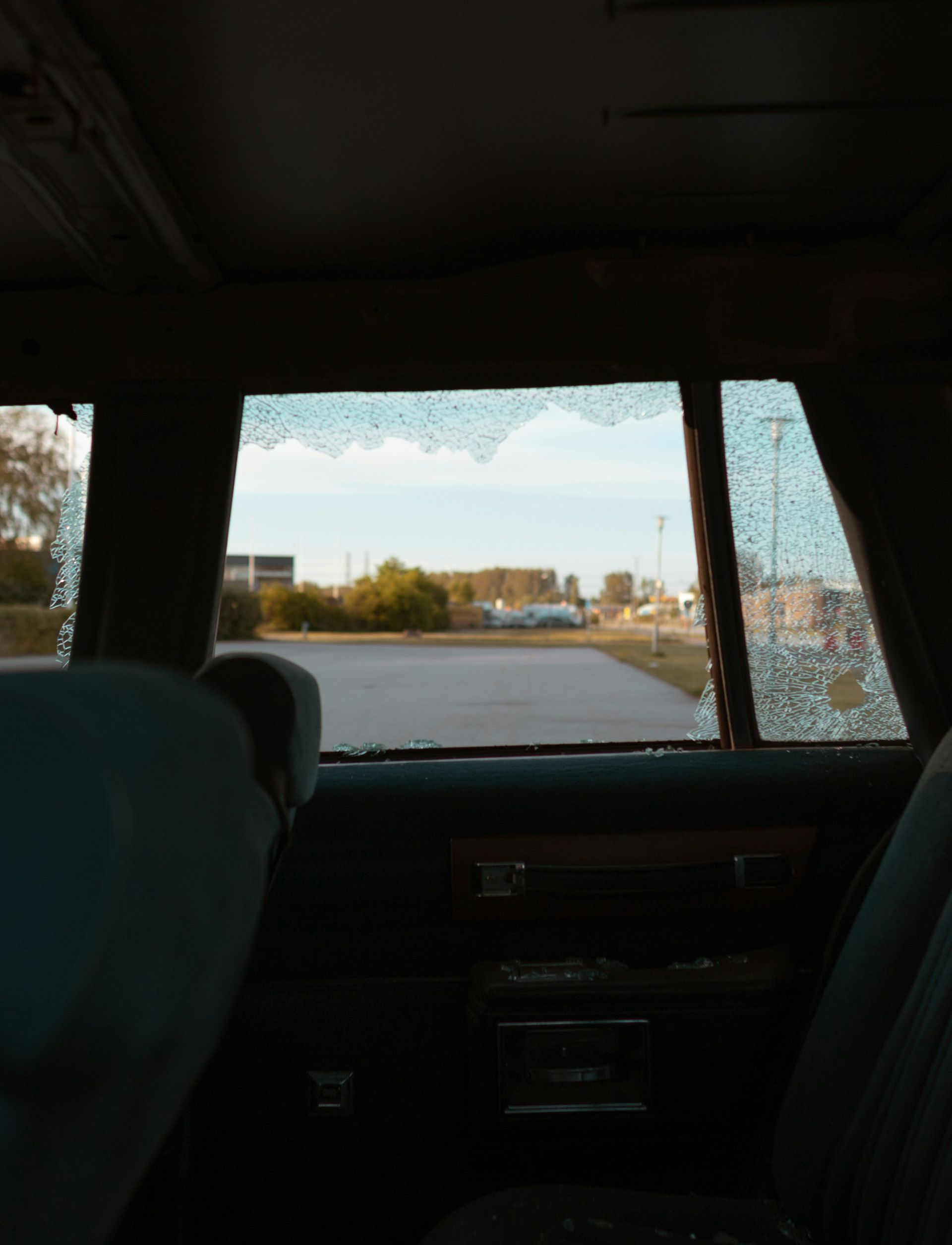 View from the back seat of a car looking out a side window with shattered glass, showing a paved parking lot and trees.