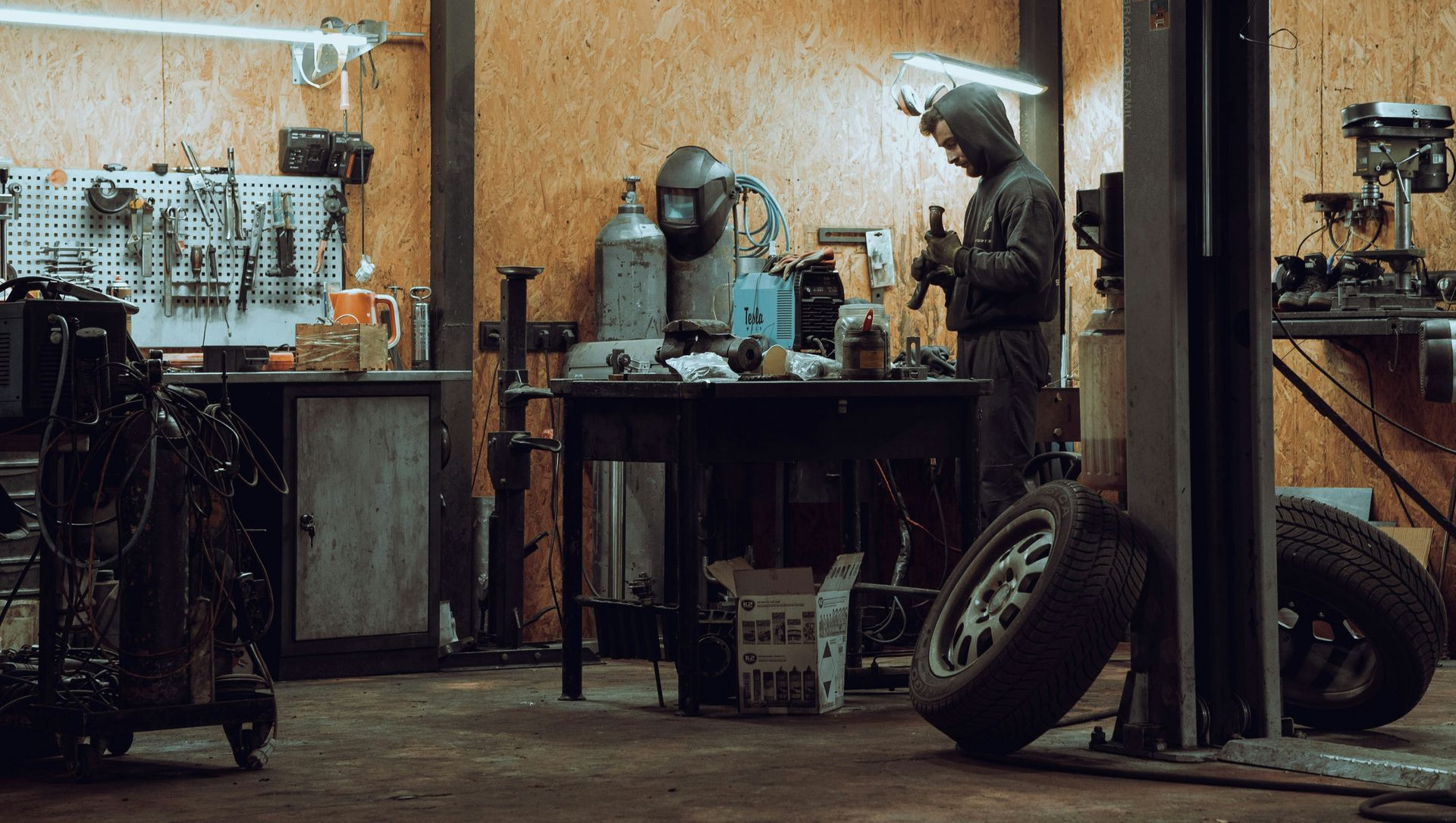 A person wearing a hooded jacket stands at a workbench in a workshop, working with equipment near tools and a tire.
