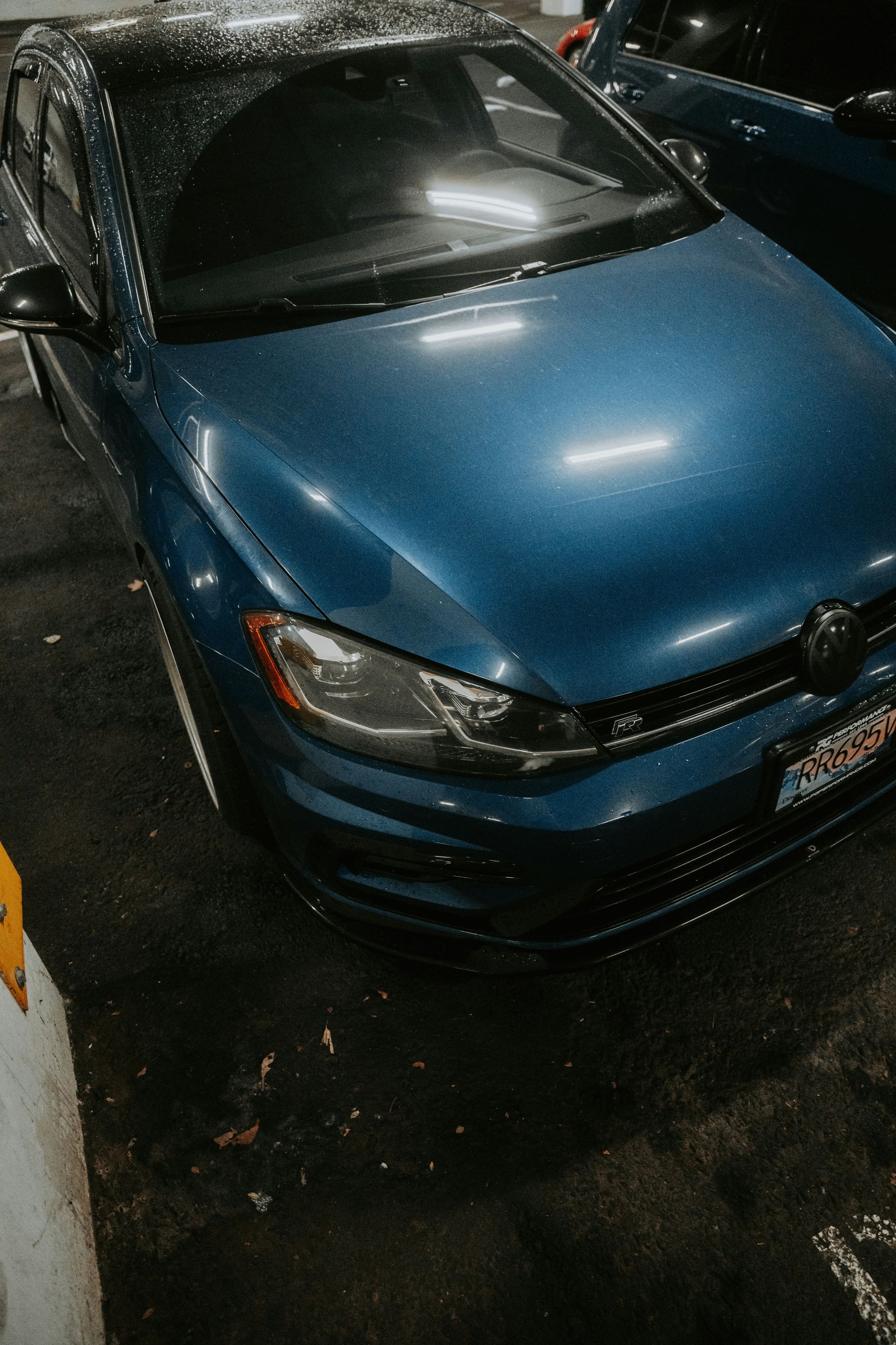 Blue Volkswagen Golf parked in a dark, wet parking lot. Headlights on, rain glistening on the car's surface.