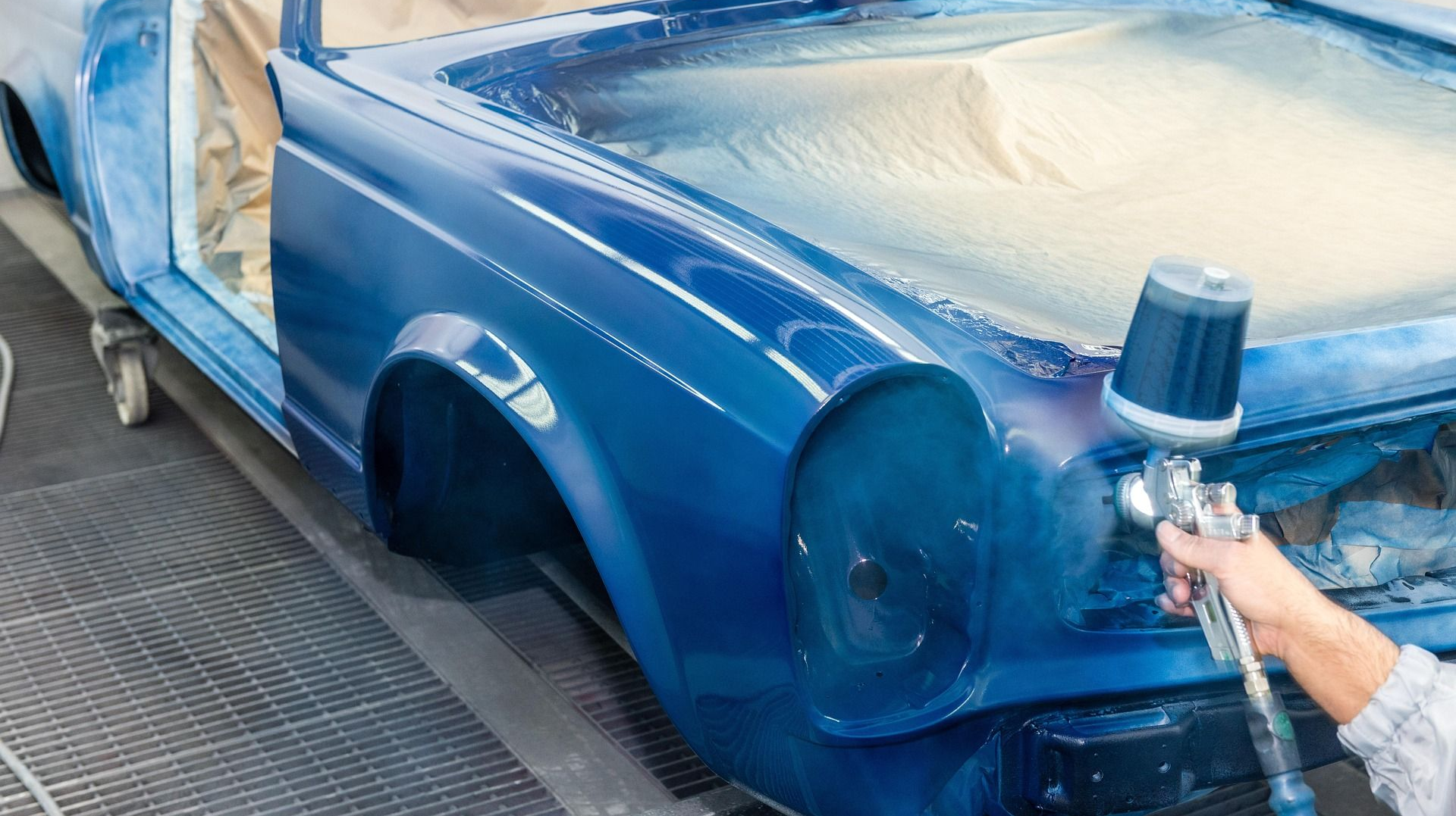 Blue convertible car being repainted; spray gun held by hand in a workshop.