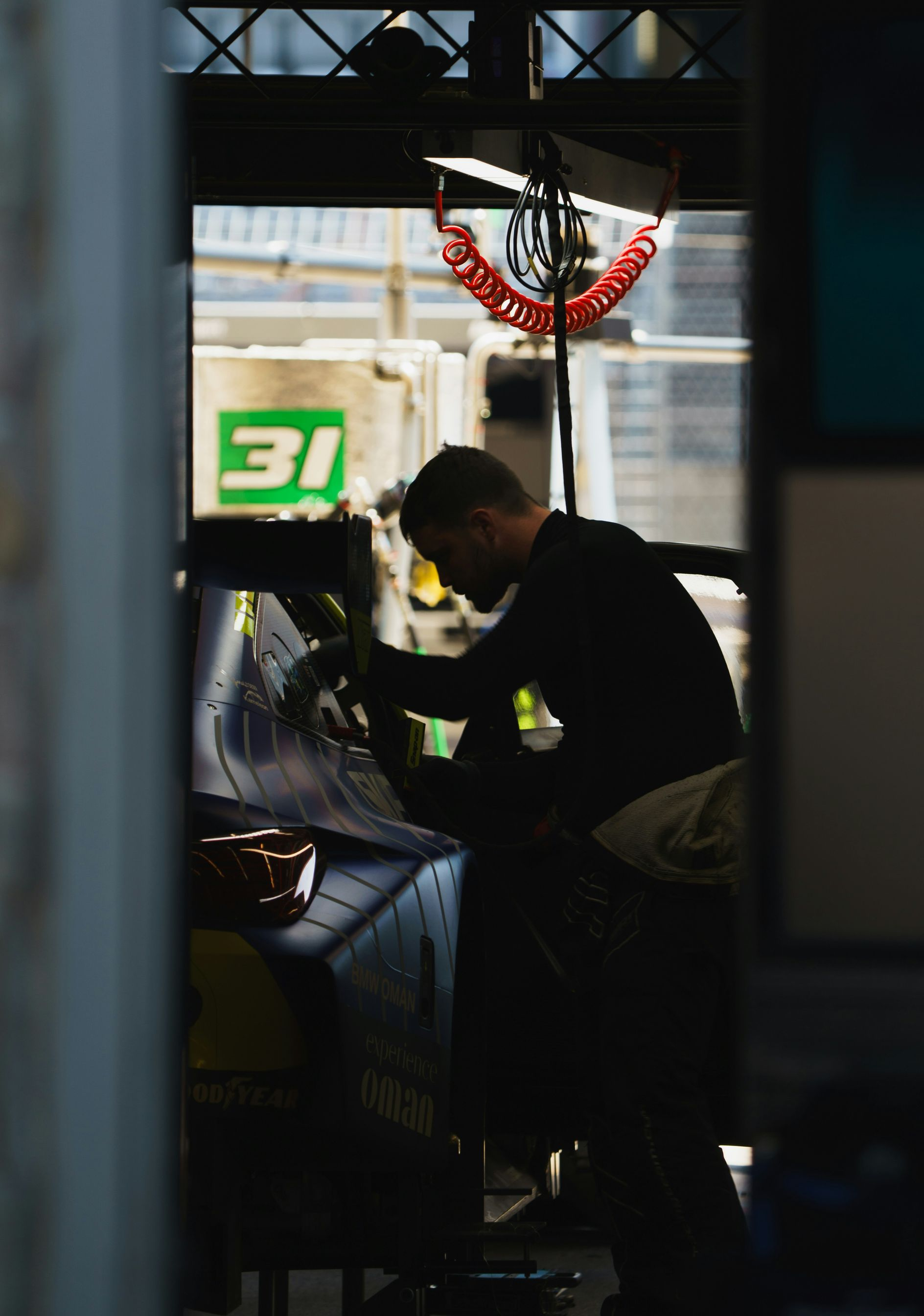 A mechanic works on a race car in a dark garage, silhouetted against a bright background with a green number 31 sign.
