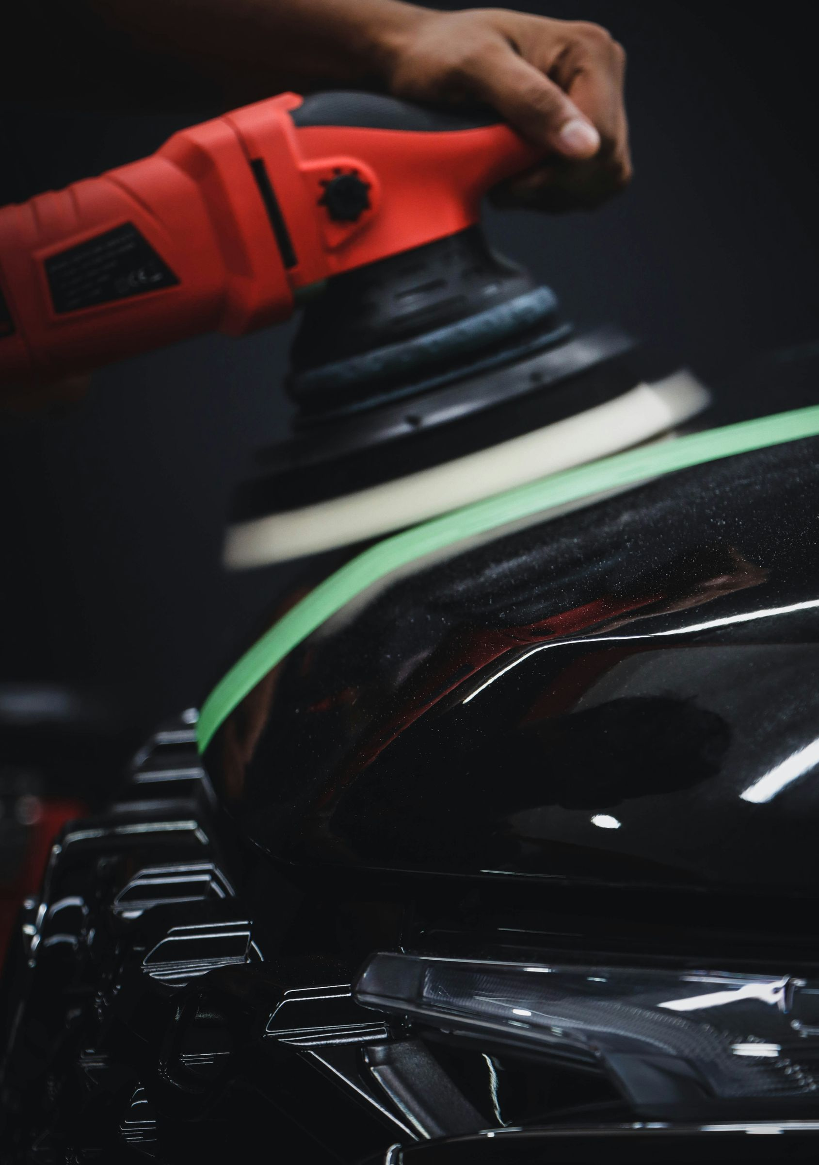 A close-up of a red orbital polisher machine being used to detail the black hood of a car with a green masking tape edge.