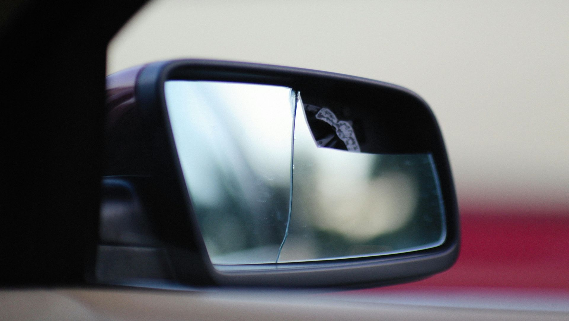 A car's side-view mirror with a vertical crack across the glass, reflecting a blurred outdoor scene.